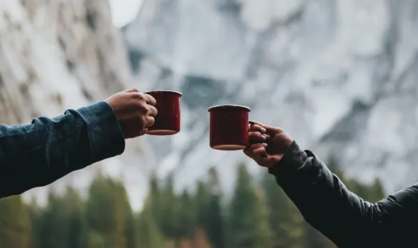 山岳風景を背景に、赤いマグカップを持って乾杯する二人の手元