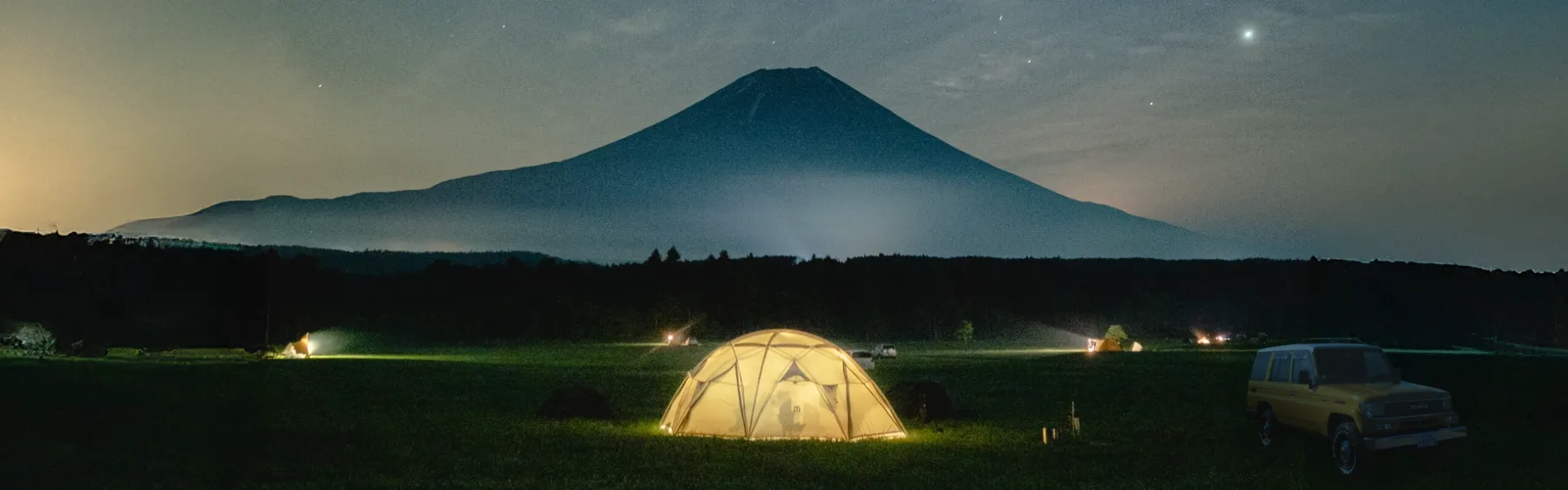 夜空の下、富士山を背景に光るテントと停車中の黄色いSUVが写るキャンプ風景