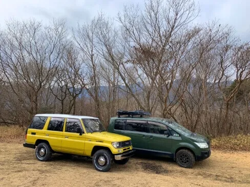 黄と緑の四輪駆動車が冬枯れの林の前に並んで停車している
